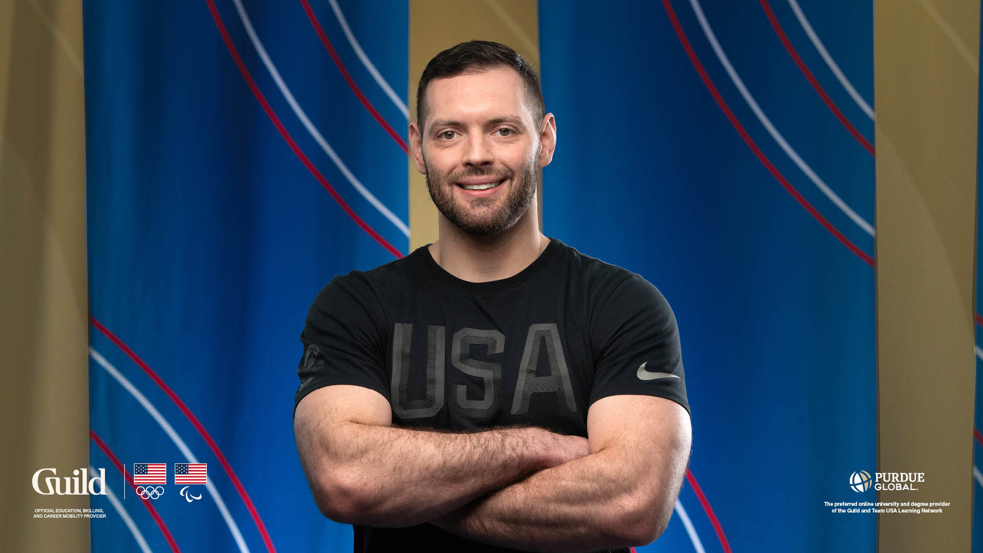 Kell Berliner poses, arms crossed, in a black Team USA shirt, in front of blue banners.
