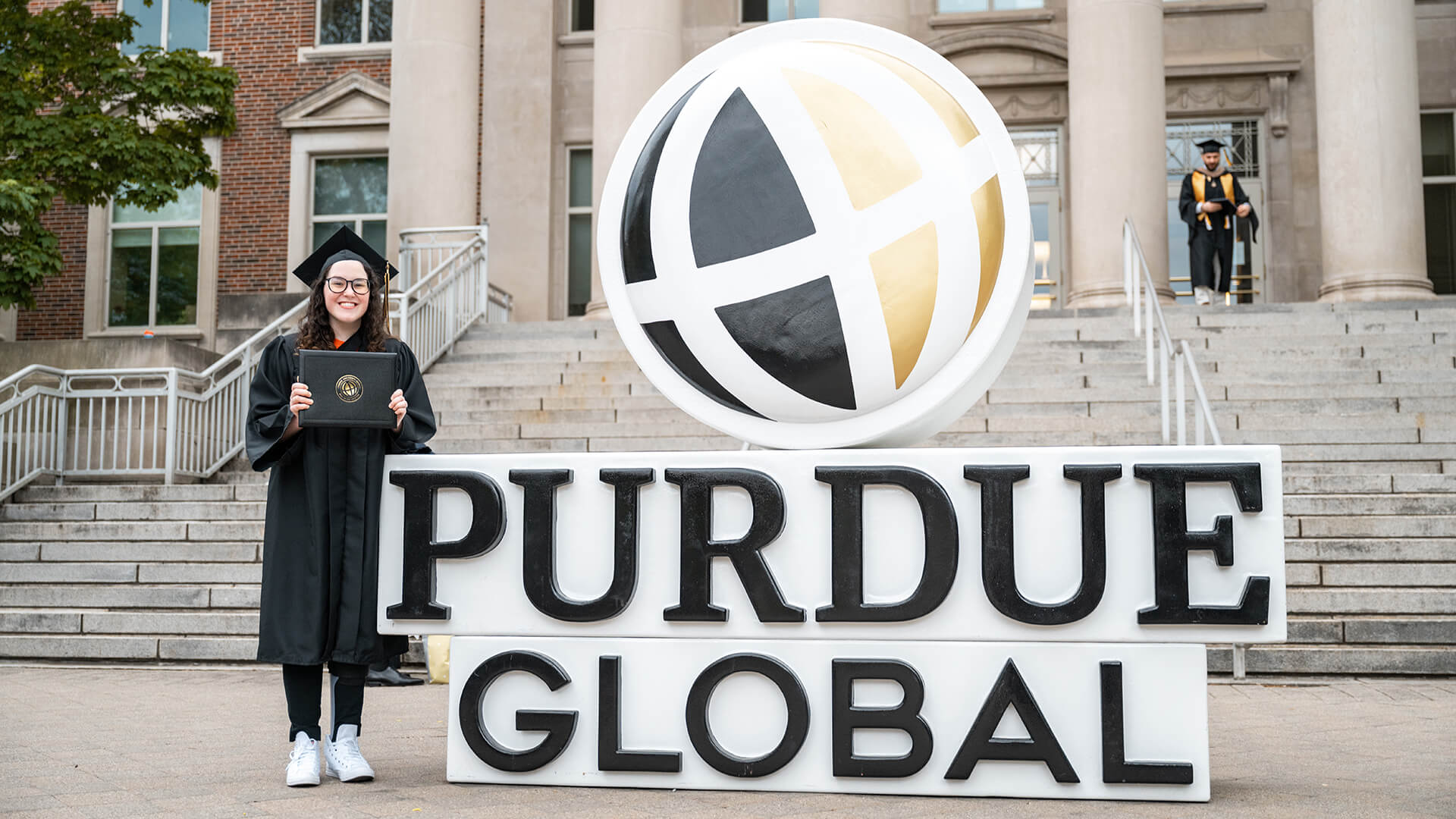 Lauryn DeLuca poses, holding her diploma and wearing a cap and gown, next to a giant Purdue Global sign outside Hovde Hall.