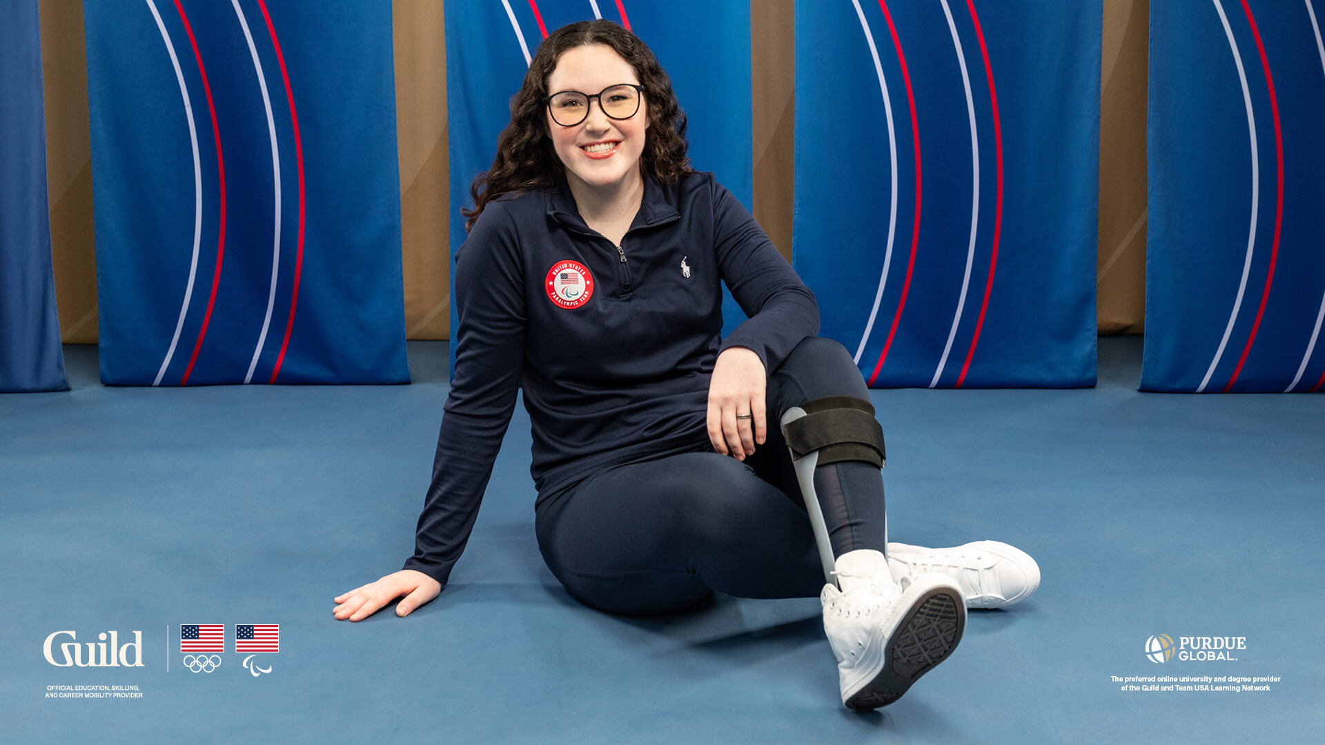 Lauryn DeLuca sits cross-legged in front of a blue banner background, a leg brace visible on her left leg.
