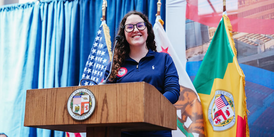 DeLuca, wearing a blue Team USA jacket, stands at a podium, both the United States and the Los Angeles flags behind her.