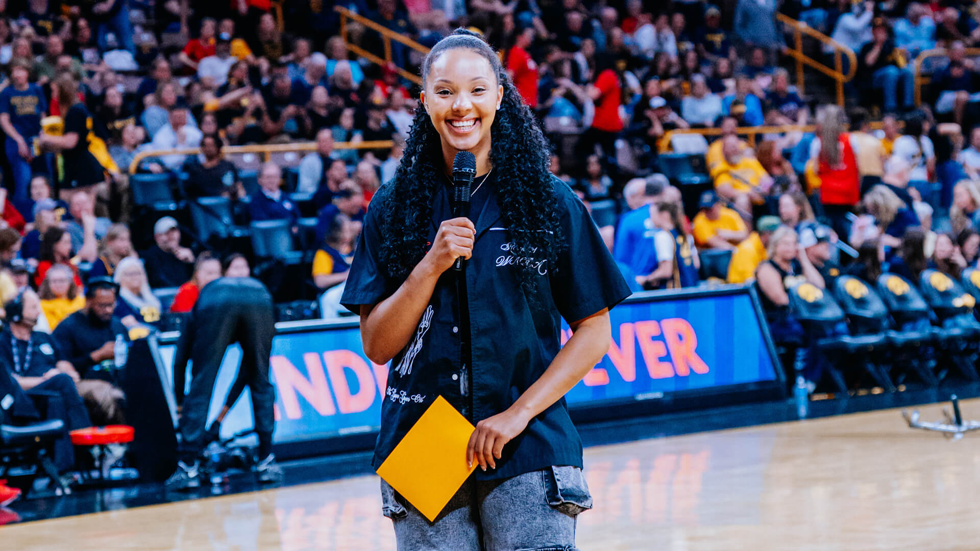Olivia West poses in Gainbridge Fieldhouse holding a mic and smiling into the camera.