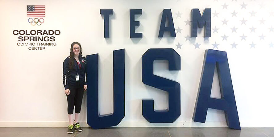 Lauryn DeLuca poses on the left side of a giant “Team USA” sign on the wall.