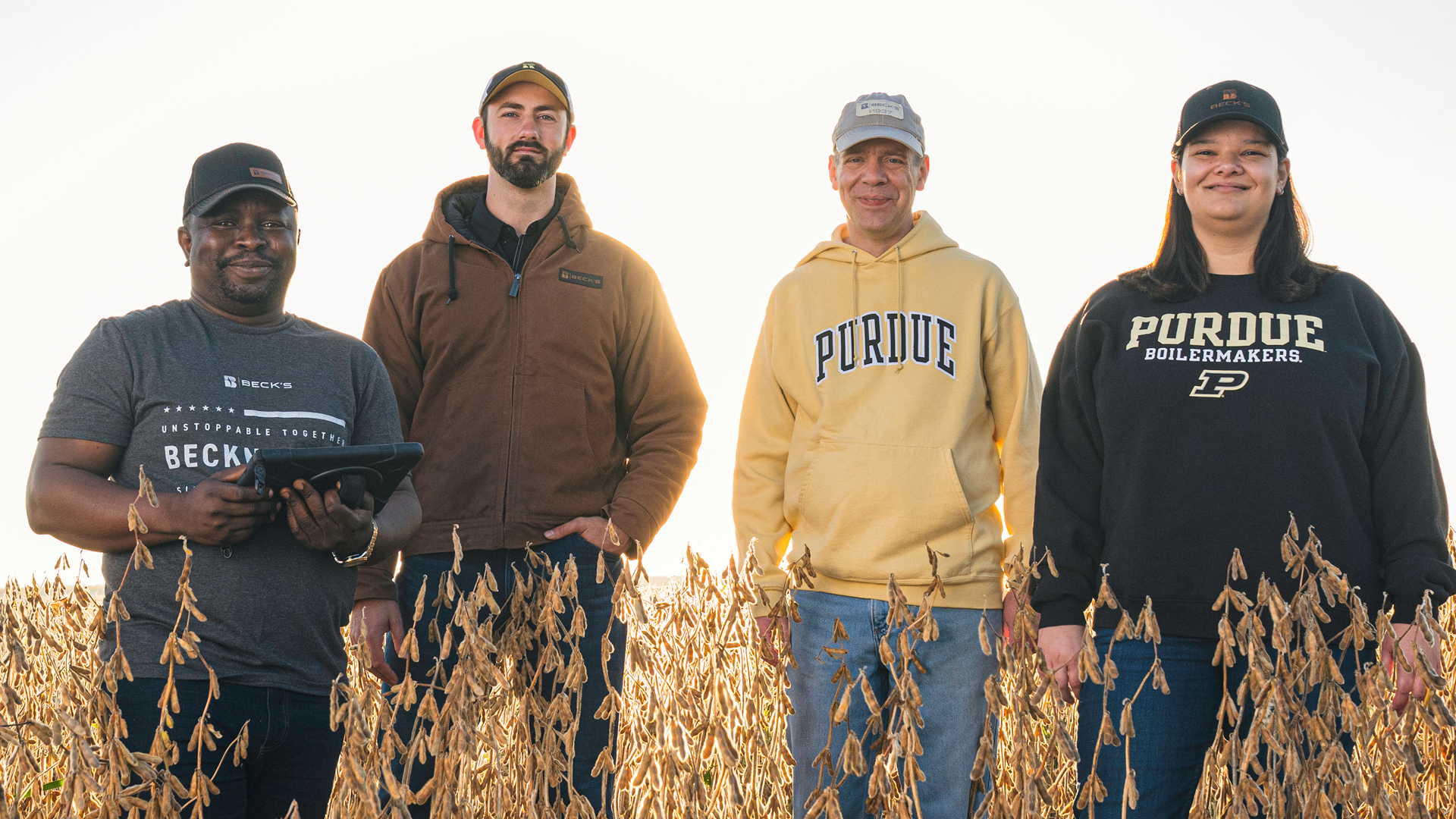 Three men and one woman standing in a field wearing Purdue and Beck’s gear.