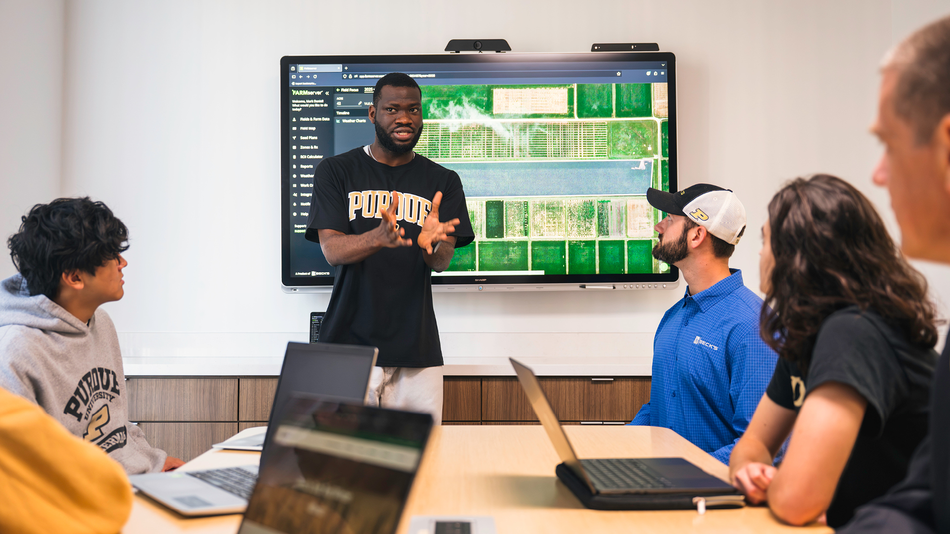 Student standing in front of digital screen presenting to a seated group of people.