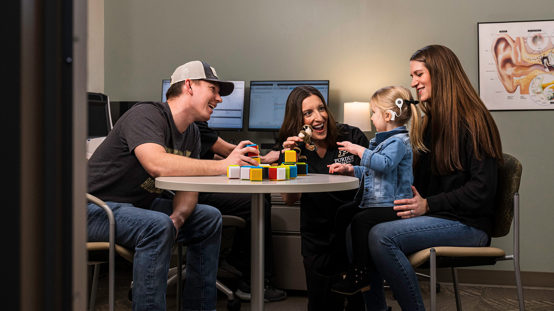 Shannon Van Hyfte, in a black Purdue polo, sits and plays with 2-year-old Callie at a desk in Purdue’s cochlear implant clinic. They’re joined by Callie’s parents, Jackson (left) and Kayla (right).