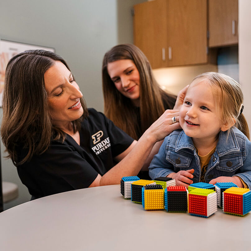 Shannon Van Hyfte, professor in Purdue’s Department of Speech, Language, and Hearing Sciences, adjusts one of Callie’s cochlear implants. Her mother, Kayla Cochran, watches from behind.