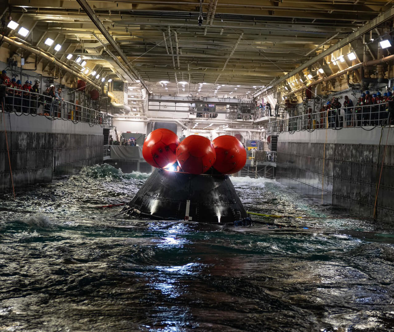 The Artemis crew module floats into the well deck of a Navy ship during a training session