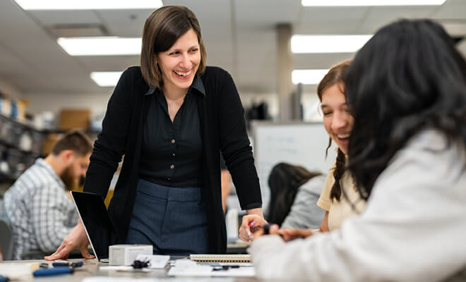 Sharon Miller and students smile while working on a project with students seated at a desk.