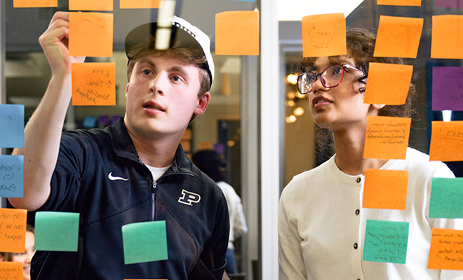 A male Purdue student and female Purdue student place colorful sticky notes on a clear glass board.